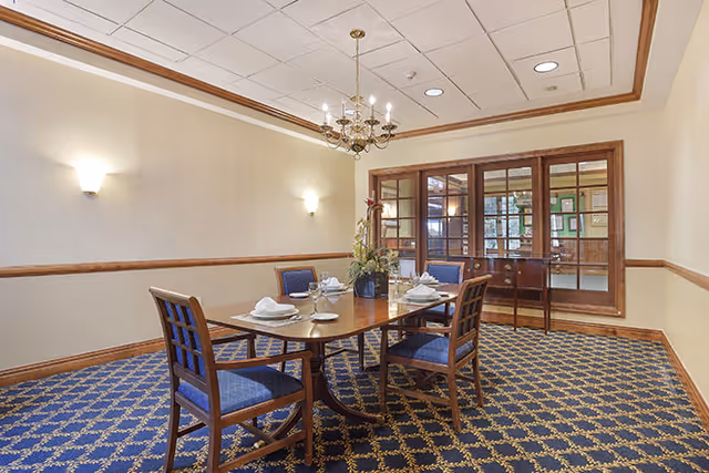 A dining room with a rectangular wooden table set for four with white napkins and plates. Four wooden chairs with blue cushioned seats surround the table. The room has a blue patterned carpet, cream-colored walls with wooden trim, two wall sconces, a chandelier, and a large window with wooden frames looking into another room.