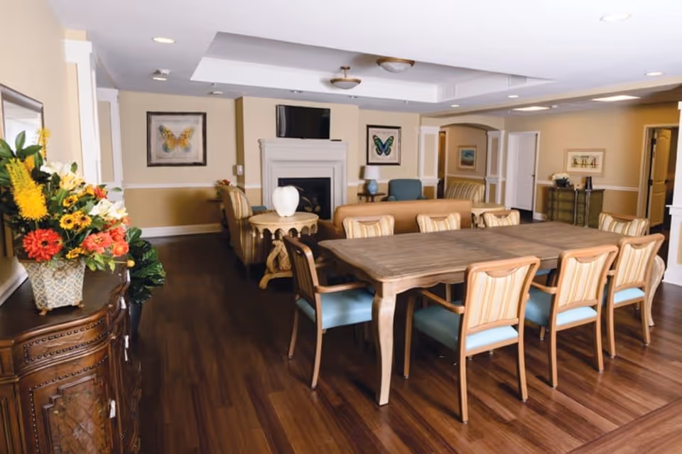 A spacious senior living common area with a large wooden table surrounded by eight chairs with striped upholstery. In the background, there is a seating area with sofas and armchairs, a fireplace with a TV mounted above it, and framed butterfly artwork on the walls. The room has wood flooring, beige walls with white trim, and recessed lighting. A decorative sideboard with a colorful flower arrangement is visible on the left side.