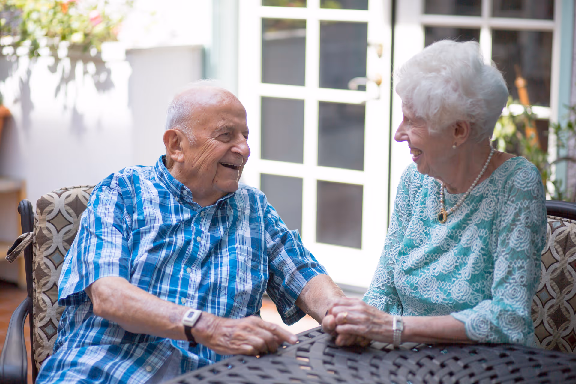 An elderly man and woman sitting at a round outdoor table, holding hands and smiling at each other. The man is wearing a blue plaid shirt and the woman is wearing a light green lace top with a pearl necklace. They are seated on cushioned chairs with a background of glass doors and some plants.