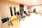A rehabilitation or physical therapy room with exercise equipment including stationary bikes and other fitness machines, a yellow chair, and a wall-mounted TV. The room has light-colored walls and flooring, with some therapy tools and posters on the walls.