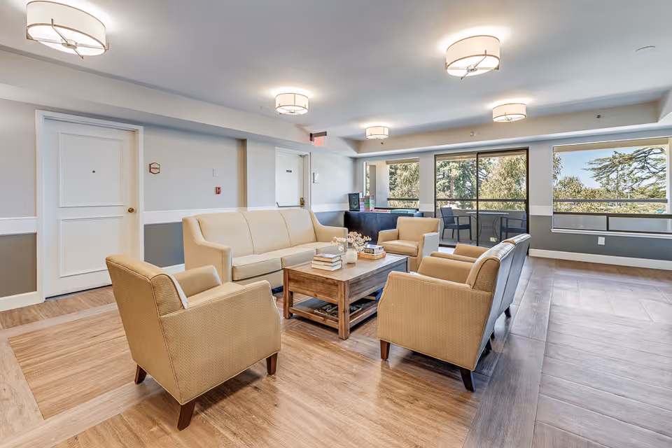Bright common lounge with beige sofas and armchairs arranged around a wooden coffee table, large windows overlooking trees, and ceiling lights.