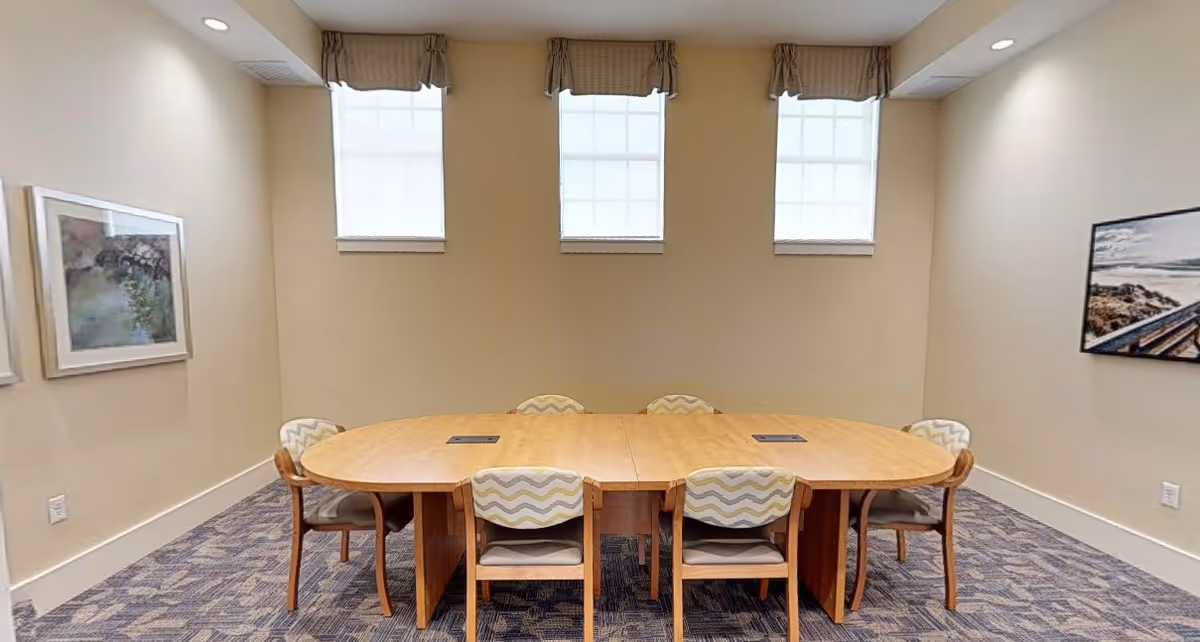 A conference or meeting room with a large oval wooden table surrounded by six chairs with patterned upholstery. The room has three tall windows with valances, beige walls, carpeted floor, and framed artwork on the walls.