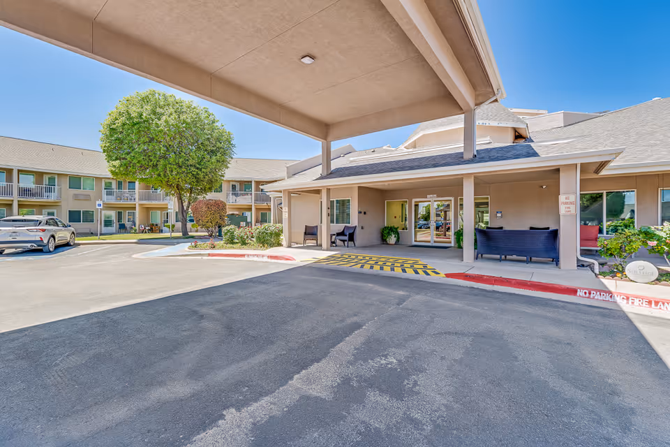 Entrance area of Solstice Senior Living at Rio Norte showing a covered drop-off zone with seating, a tree, shrubs, and a two-story building with balconies in the background under a clear blue sky.