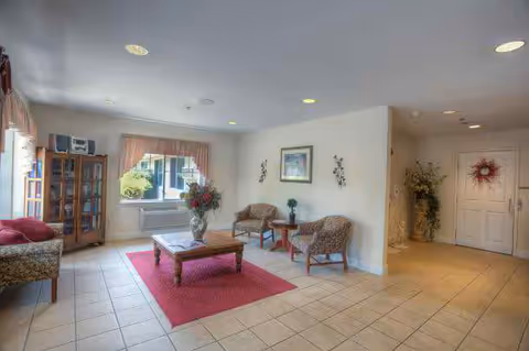 A bright and spacious living room area with tiled floors, a red area rug, a wooden coffee table with a flower vase, two upholstered chairs with a small side table, a cushioned bench, a wooden cabinet with a stereo system, and a window with curtains letting in natural light. There is also a hallway with a door decorated with a wreath and a plant in the corner.