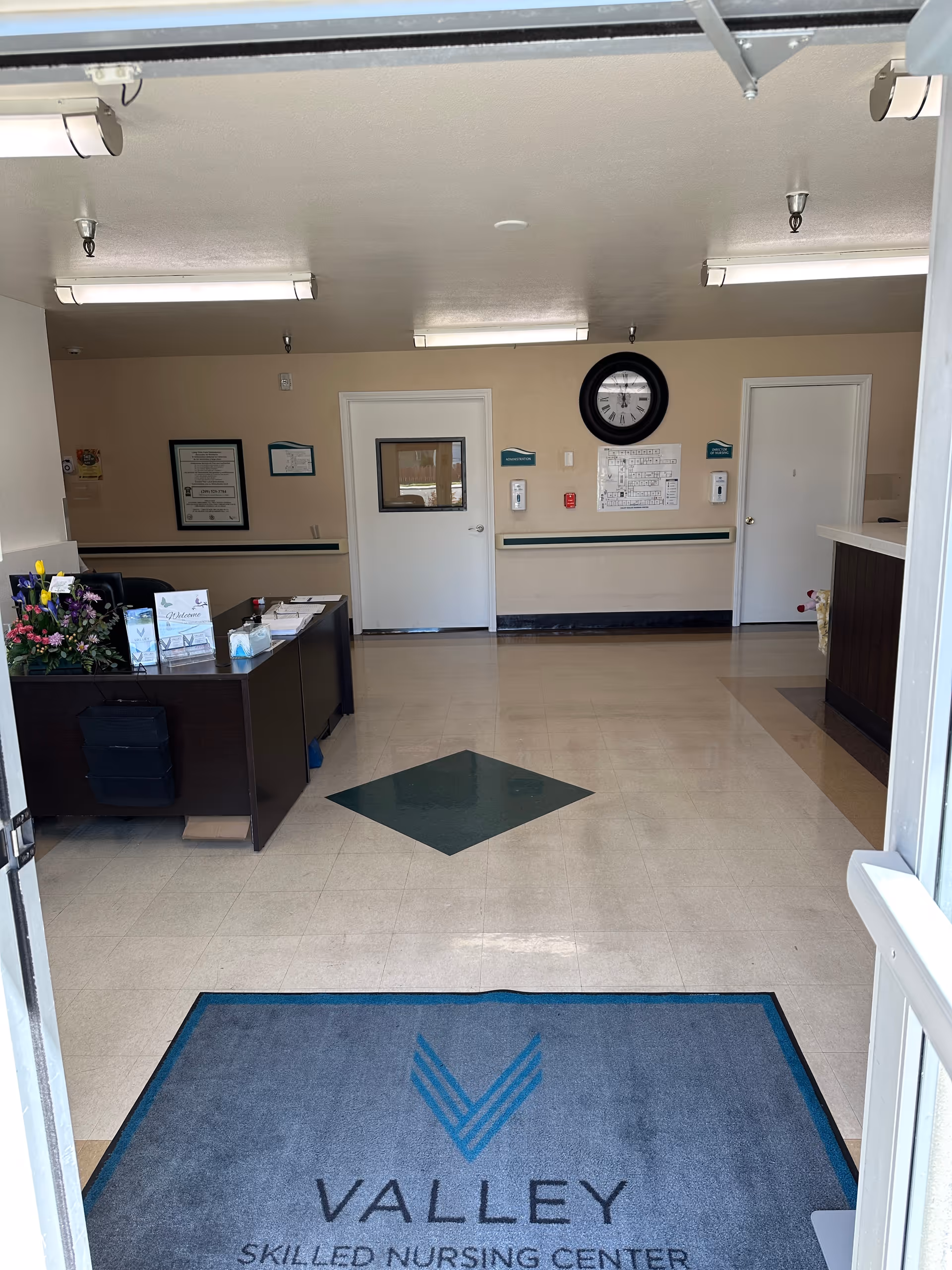 Interior entry lobby of a nursing facility with a reception desk, wall clock, two doors, and a floor mat reading 'Valley Skilled Nursing Center'.