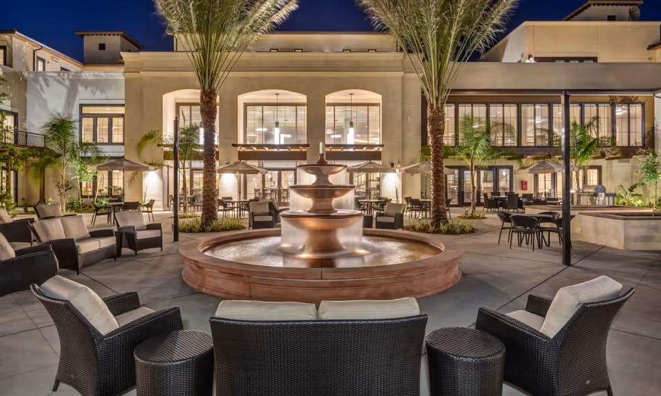 Outdoor courtyard area at night with a large tiered water fountain in the center, surrounded by cushioned wicker chairs and tables. Palm trees and patio umbrellas are visible, with a multi-story building featuring large windows in the background.