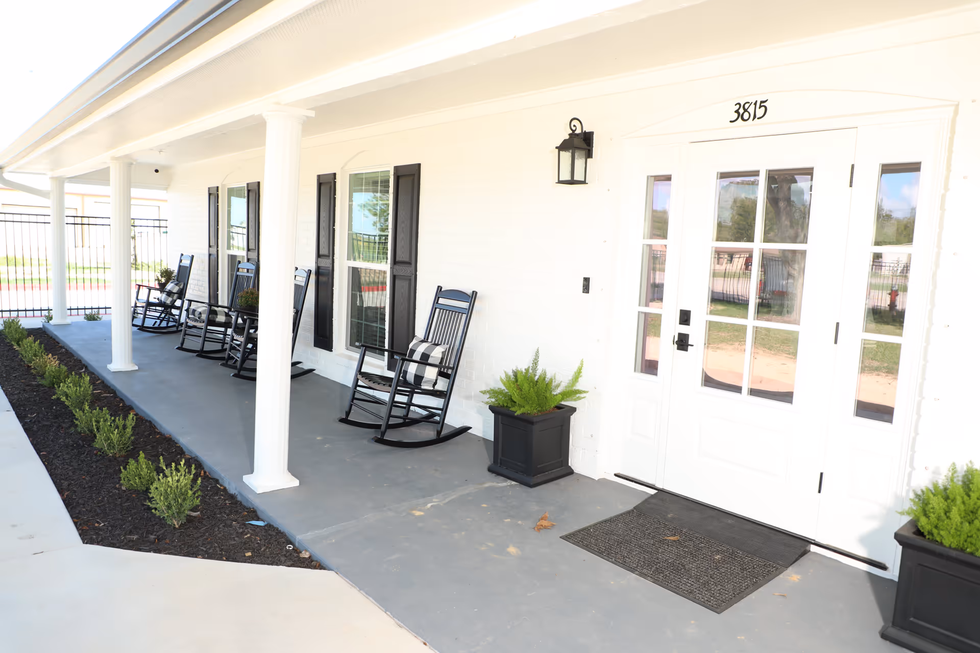 Front porch of a building with white brick walls, black rocking chairs with cushions, black window shutters, two black planters with green plants, white columns supporting the roof, and a white door with glass panels and the number 3815 above it.