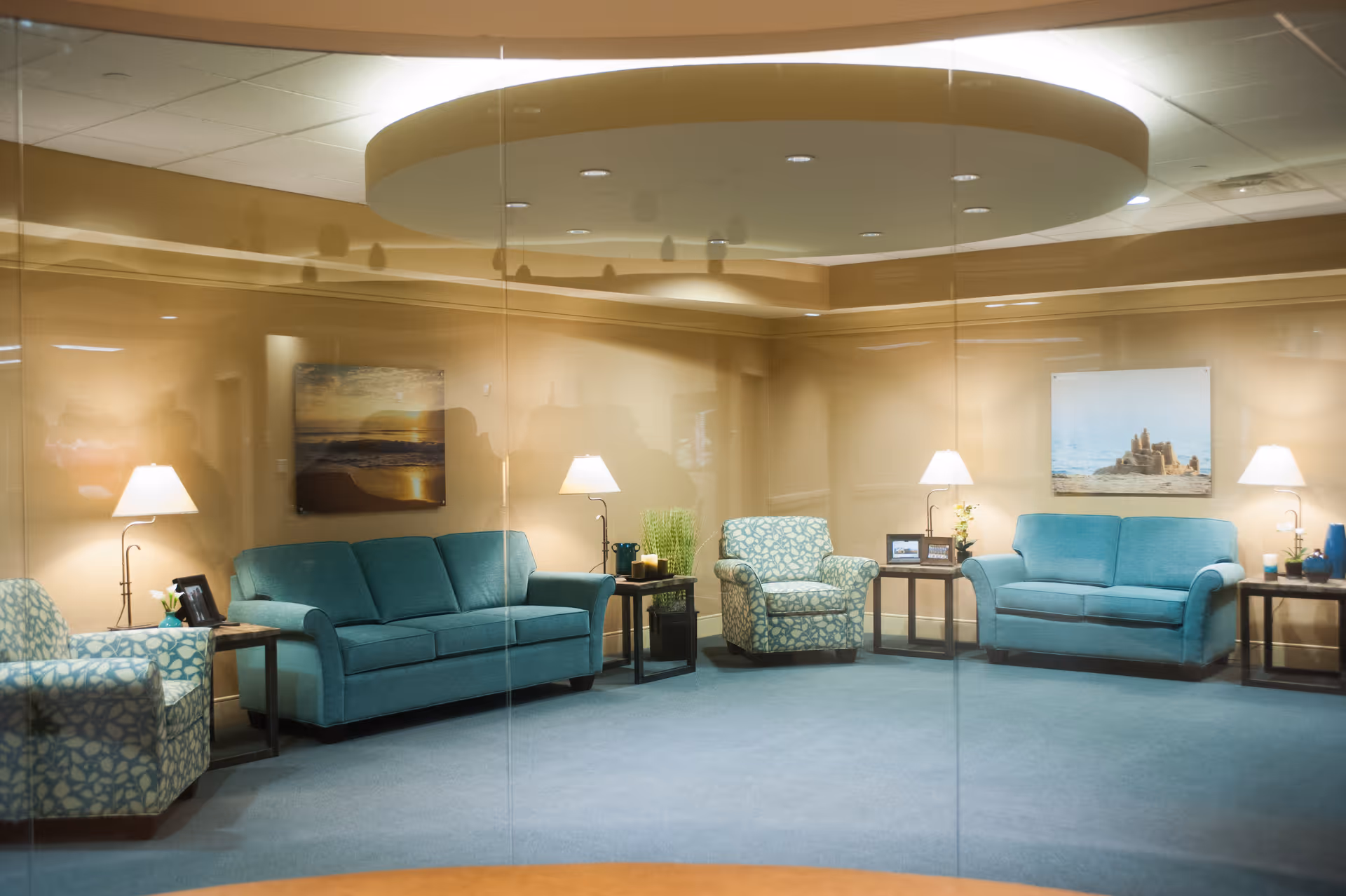 A cozy living room area with teal and patterned armchairs and sofas arranged around small side tables with lamps. The walls are beige with framed pictures, and the ceiling has a circular recessed lighting feature.