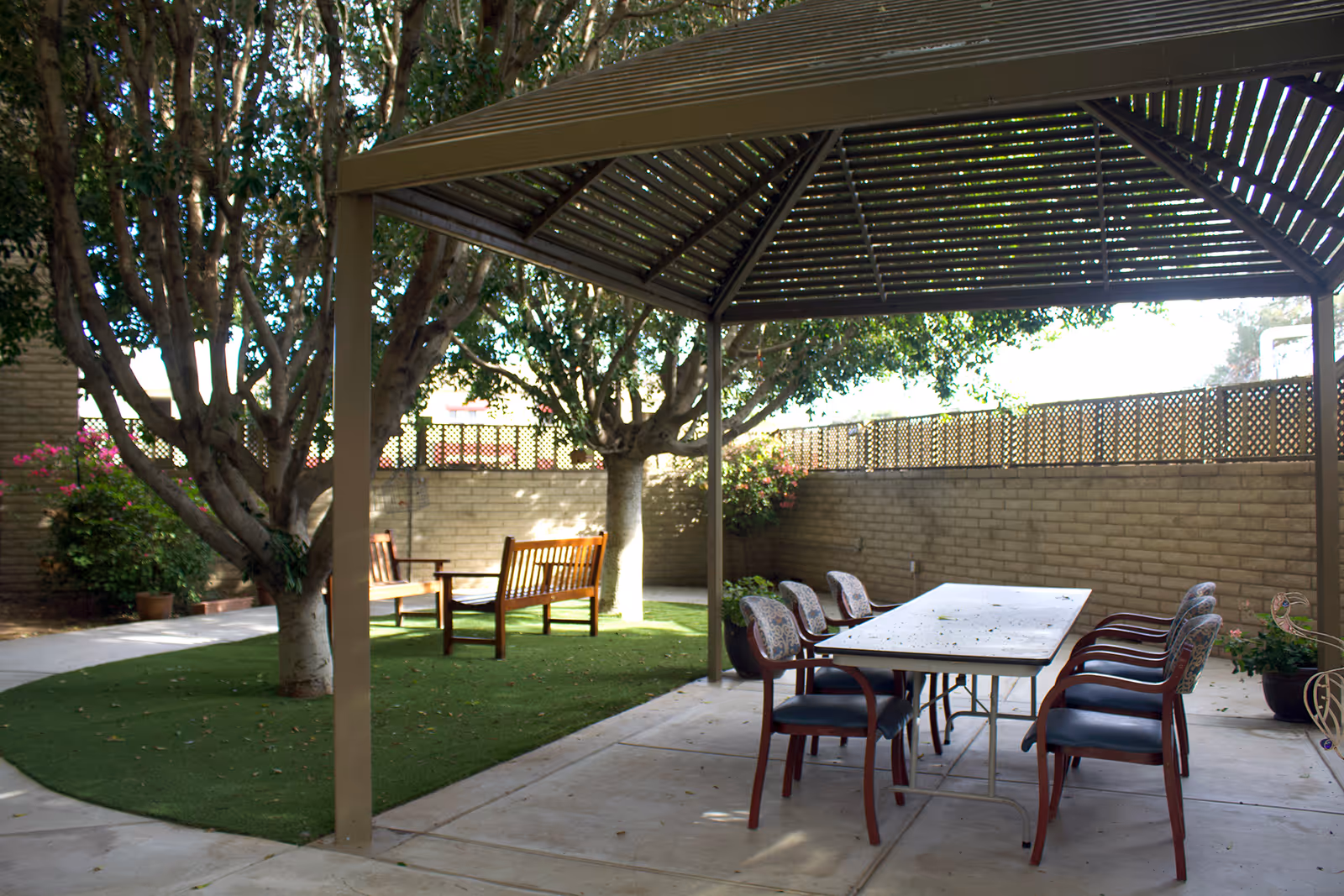 Outdoor patio area with a metal pergola covering a rectangular table surrounded by eight chairs. There are trees and green artificial grass nearby, along with wooden benches and a brick wall enclosing the space.