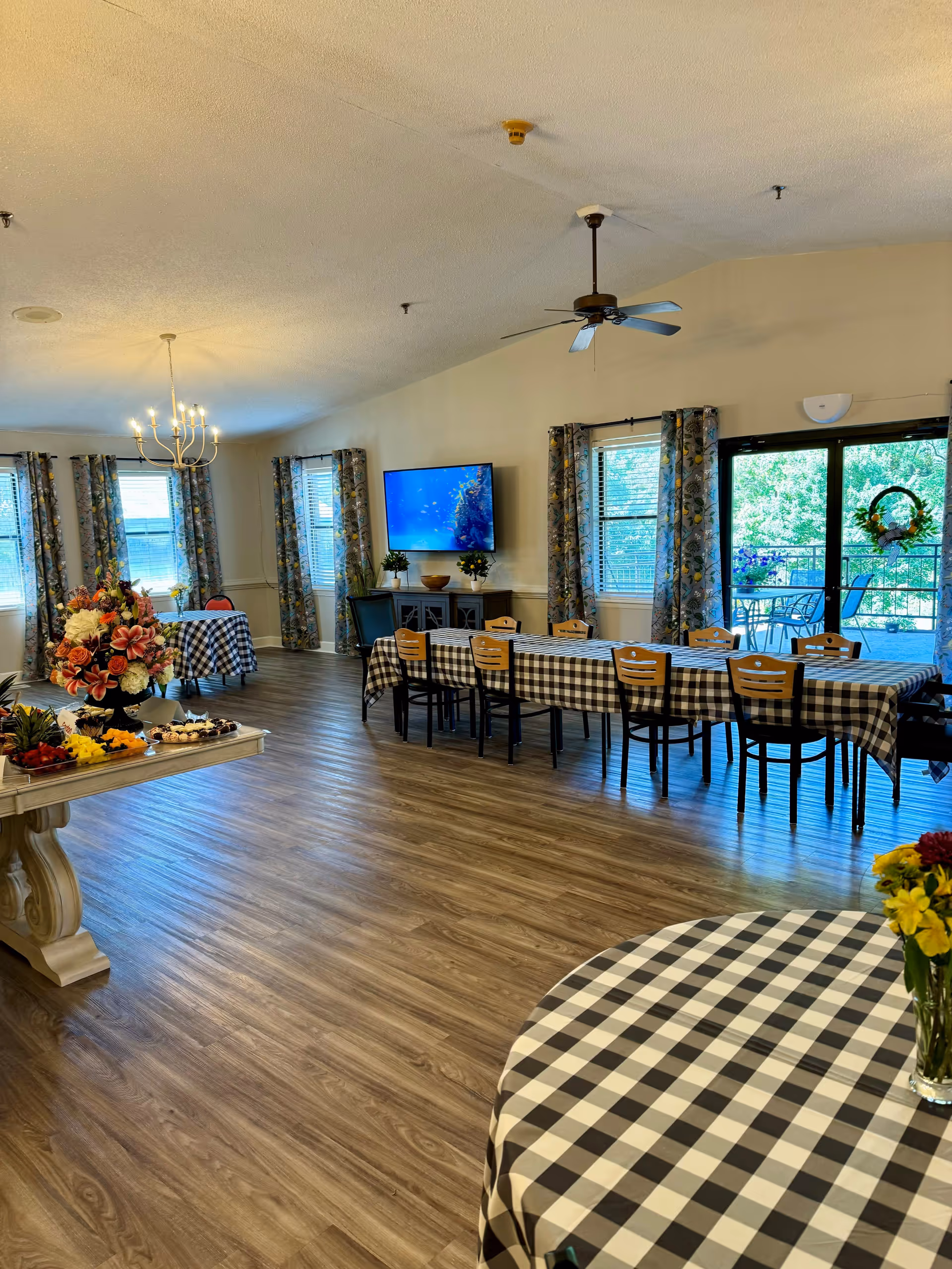 A spacious dining area in Silver Lakes Senior Living with multiple tables covered in black and white checkered tablecloths. There are chairs around the tables, a chandelier hanging from the ceiling, a ceiling fan, and a large flat-screen TV mounted on the wall. The room has large windows with patterned curtains and a glass door leading to an outdoor patio with seating. A table with a floral arrangement and fruit platters is also visible.