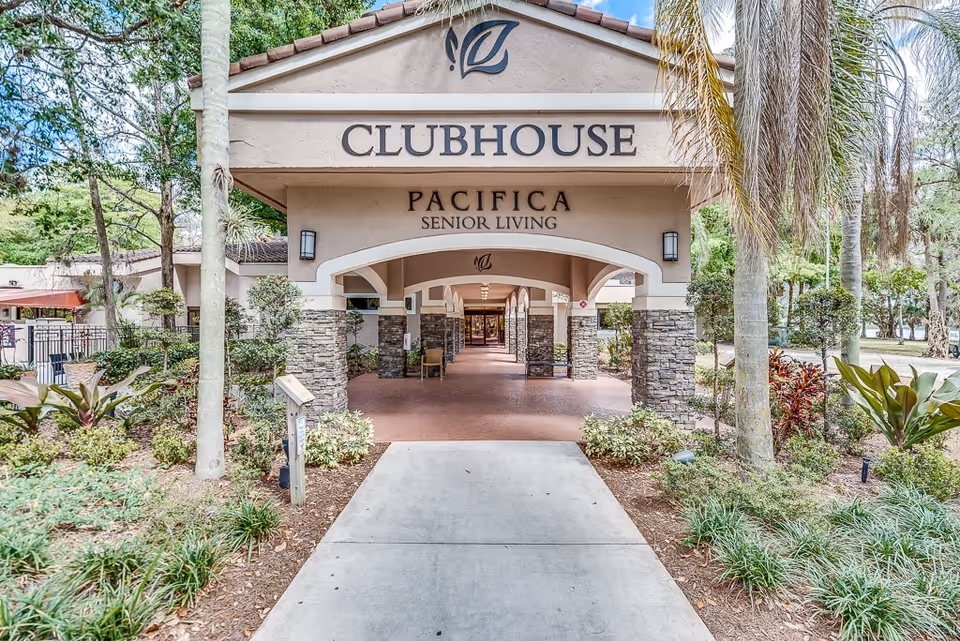 Entrance to a clubhouse at a senior living facility named Pacifica Senior Living, featuring a covered walkway with stone pillars and surrounded by landscaped greenery and palm trees.