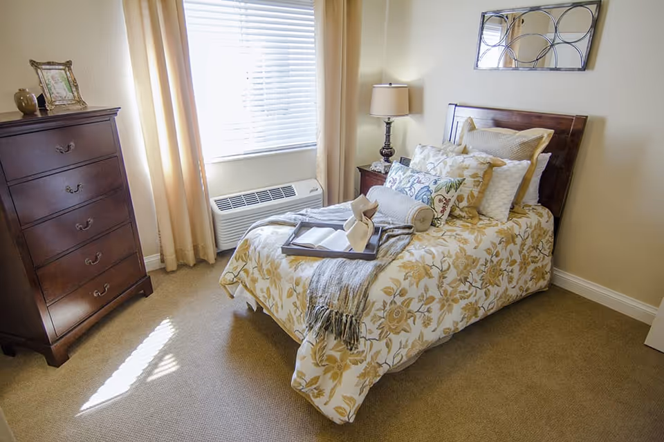 A cozy bedroom with a single bed covered in floral patterned bedding and multiple pillows. A wooden nightstand with a lamp and a wooden chest of drawers are also visible. A window with blinds and beige curtains lets in natural light.
