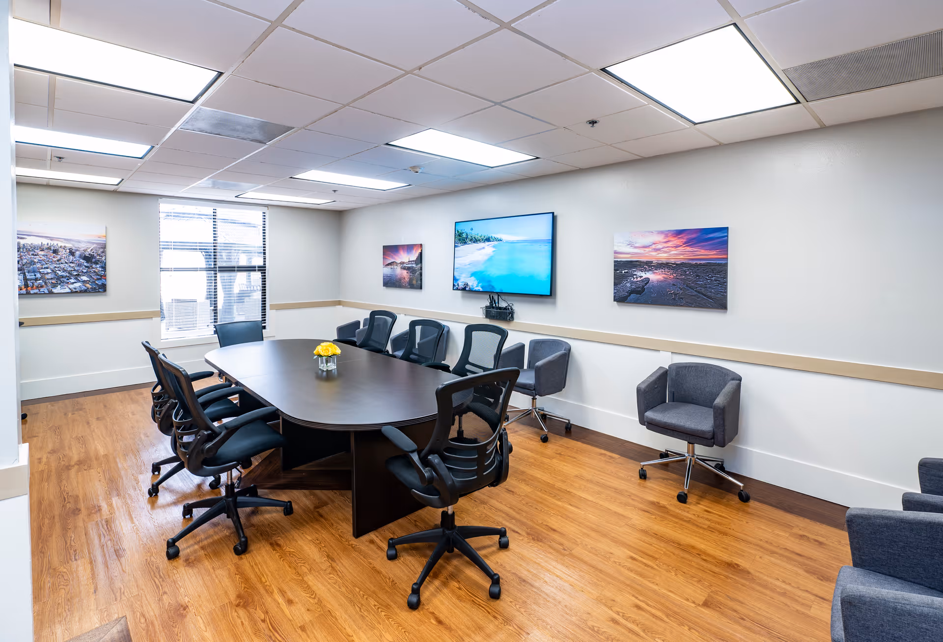 A modern conference room with a large dark wooden table surrounded by black and gray office chairs on a wooden floor. The walls are white with three colorful landscape photos and a large flat-screen TV mounted on one wall. A small vase with yellow flowers is placed in the center of the table. The room has a window with blinds and a white ceiling with fluorescent lighting.
