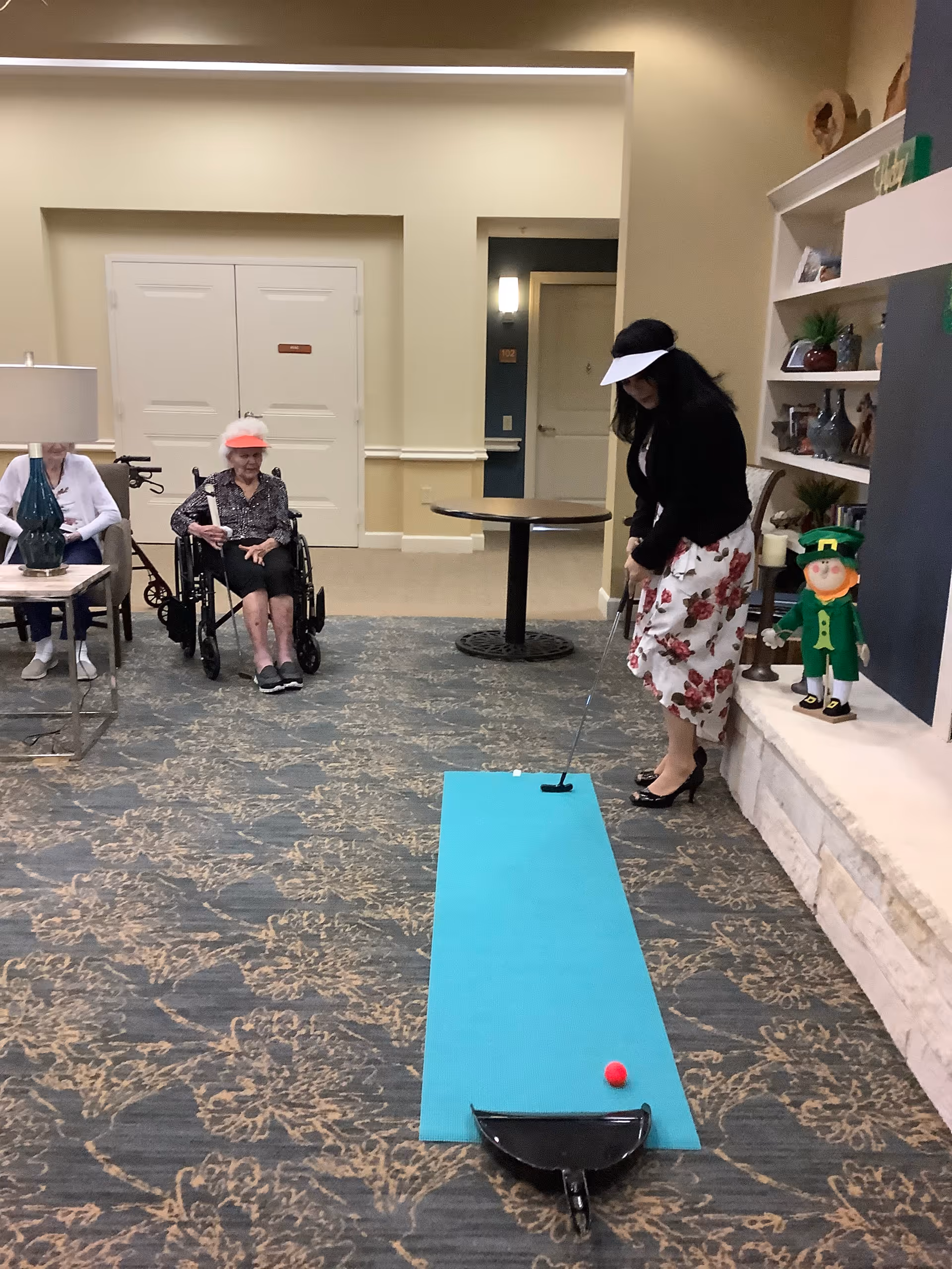 Residents in a senior living common room watch a woman putting on a small indoor putting mat.