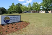 Outdoor view of a healthcare facility with a large grassy area, trees, and a sign in the foreground that reads 'Blue Ridge Healthcare Montrose Bay'. The building is visible in the background under a partly cloudy sky.
