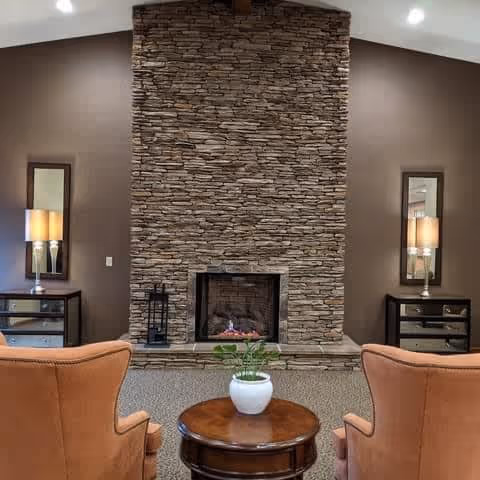 A cozy interior living area featuring a tall stone fireplace centered on a brown wall. On either side of the fireplace are matching dark wood side tables with drawers, each topped with a lamp and a rectangular mirror above. In front of the fireplace are two orange upholstered armchairs facing a round wooden coffee table with a white potted plant on it.