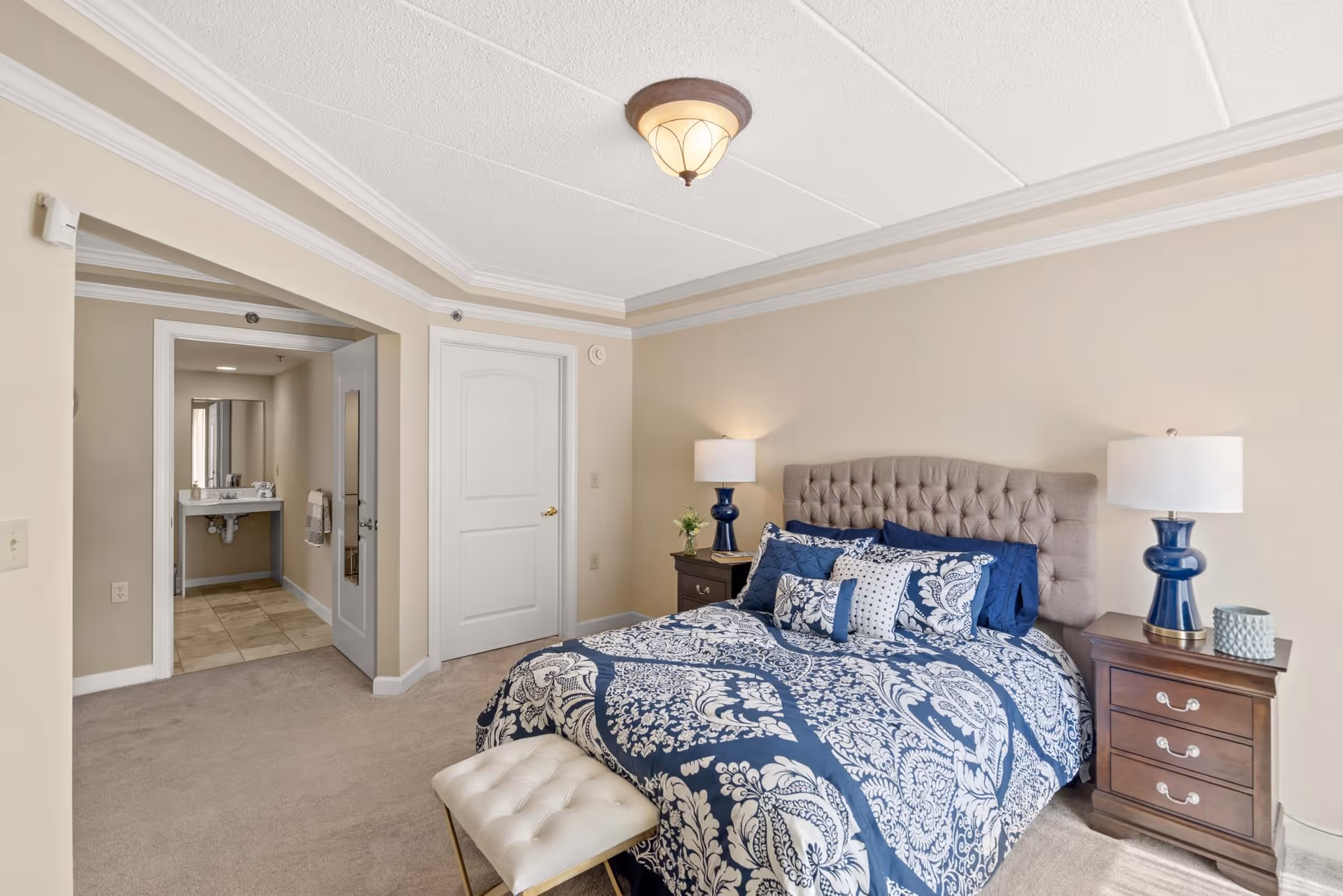 A bedroom with a large bed featuring a blue and white patterned comforter and multiple pillows. The bed has a tufted headboard and is flanked by two wooden nightstands with blue lamps. There is a small upholstered bench at the foot of the bed. The room has beige walls, carpeted floor, and a ceiling light fixture. An open doorway leads to a bathroom area with a sink and mirror visible.