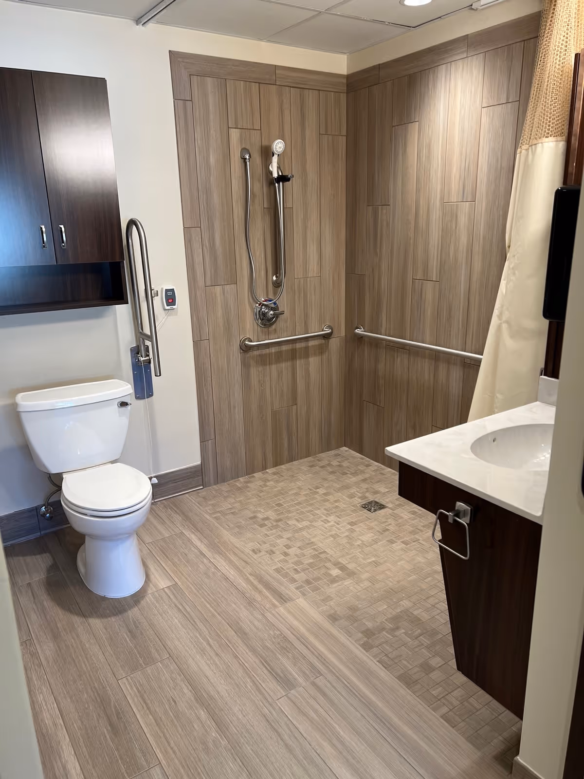A modern bathroom with wood-look tile flooring and walls. The room features a white toilet with a grab bar beside it, a walk-in shower with a handheld showerhead and two horizontal grab bars, and a sink with a white countertop and dark wood cabinetry. There is also a wall-mounted cabinet above the toilet and a beige shower curtain partially visible.