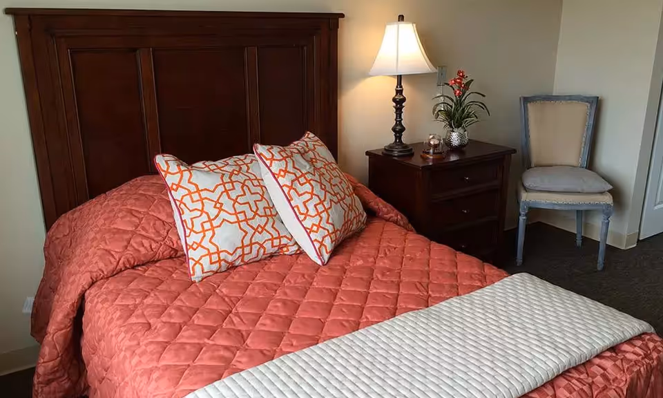 A bedroom with a wooden headboard bed covered with a coral quilted bedspread and two white pillows with orange geometric patterns. Next to the bed is a dark wooden nightstand with a lamp, a small plant, and a decorative item. A beige upholstered chair with a wooden frame is placed against the wall.