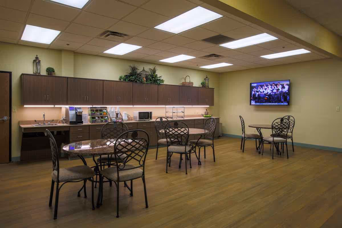 A communal dining area with several round tables and metal chairs with cushioned seats. Along one wall, there is a kitchenette with cabinets, a coffee machine, a microwave, and various supplies. A flat-screen TV is mounted on the opposite wall, displaying a group of people. The room has a wooden floor and a ceiling with fluorescent lighting.