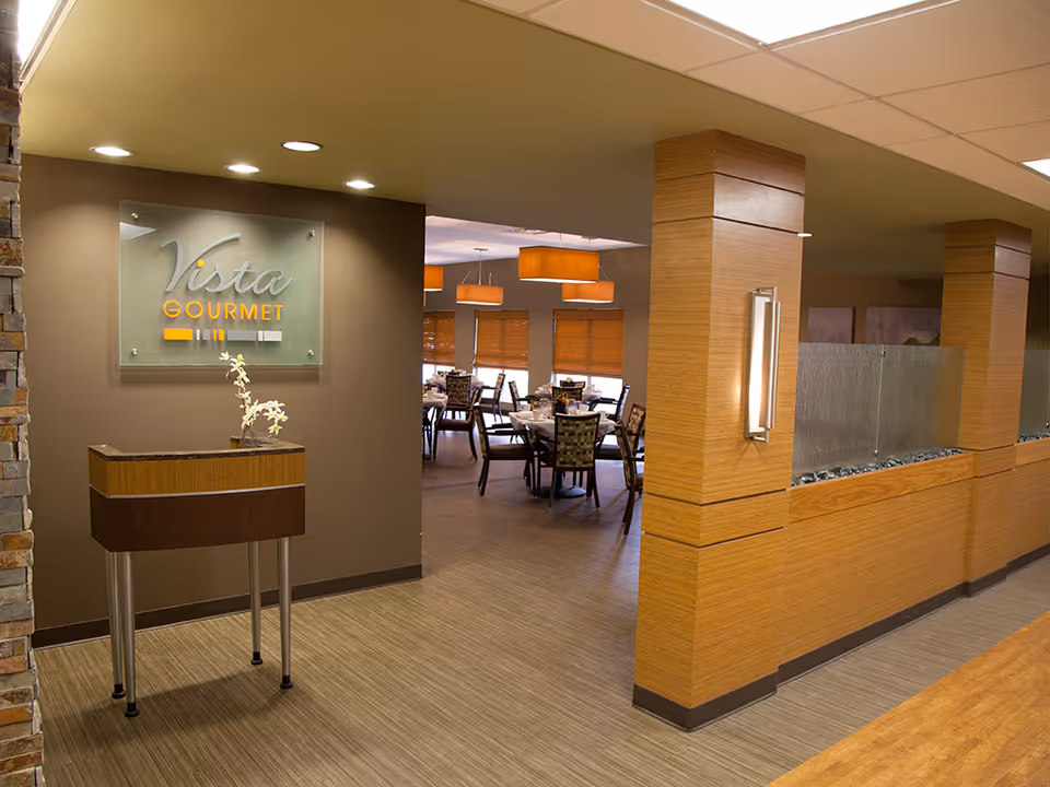 Interior view of a dining area in a senior living facility named Vista Gourmet. The image shows a modern, warmly lit space with wooden flooring and columns, a small table with a flower vase near the entrance, and several dining tables and chairs set up in the background under orange pendant lights.