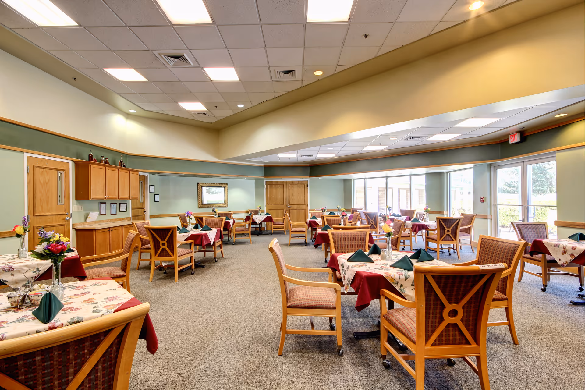 A spacious dining room with multiple tables covered in floral and burgundy tablecloths, each set with green folded napkins and small flower vases. Wooden chairs with patterned cushions surround the tables. The room has large windows letting in natural light, light green walls with wooden trim, and a ceiling with recessed lighting. There are wooden cabinets and doors along one wall.