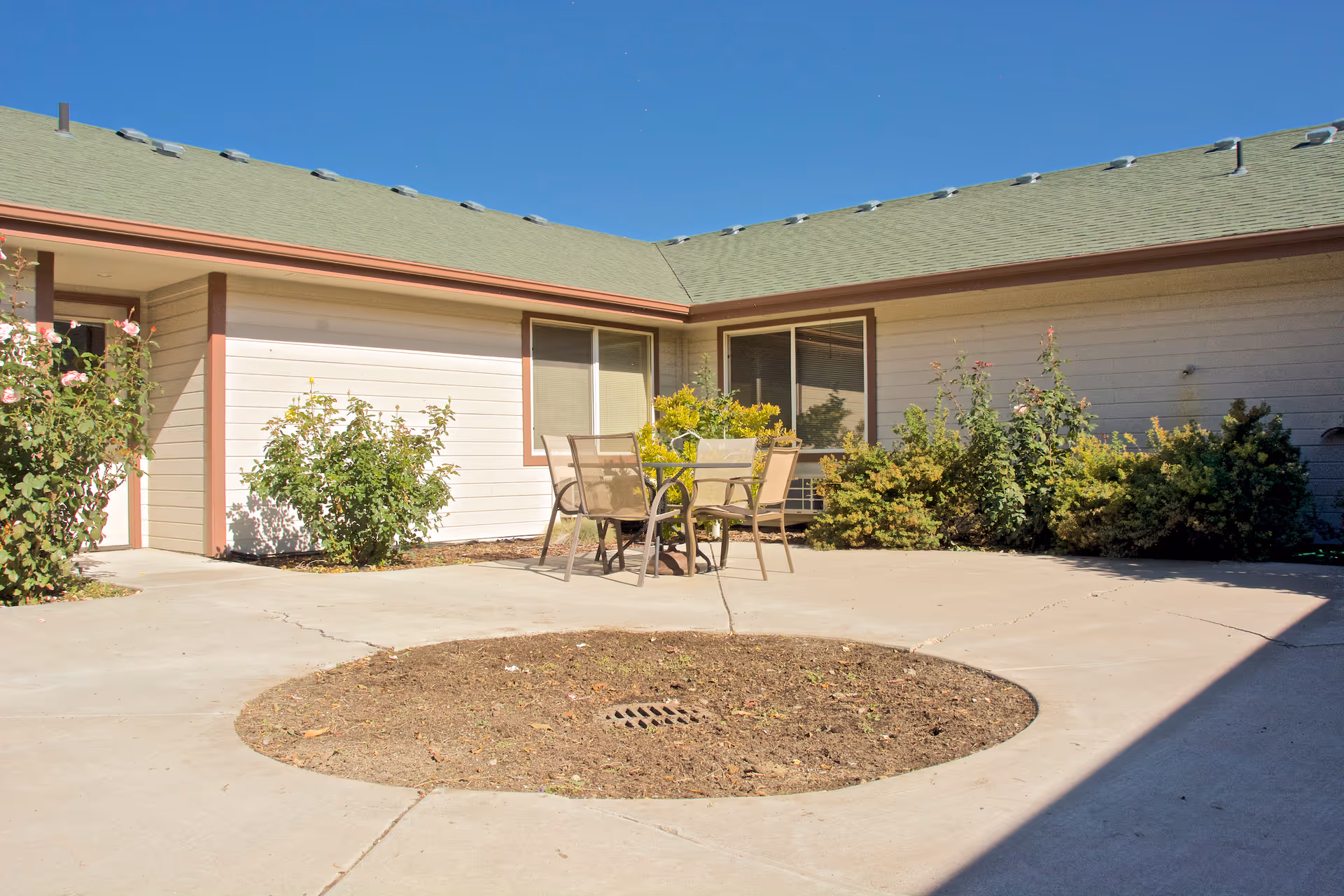 Outdoor courtyard area with a round patch of soil in the center surrounded by concrete. There is a small table with four chairs on the concrete near the building. The building has beige siding with brown trim and a green roof. Bushes and plants are along the building walls under clear blue sky.