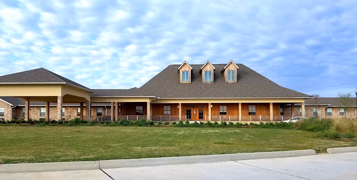 Front exterior view of LaCour House Assisted Living Community building with a large covered entrance, three dormer windows on the roof, and a well-maintained lawn in front under a partly cloudy sky.