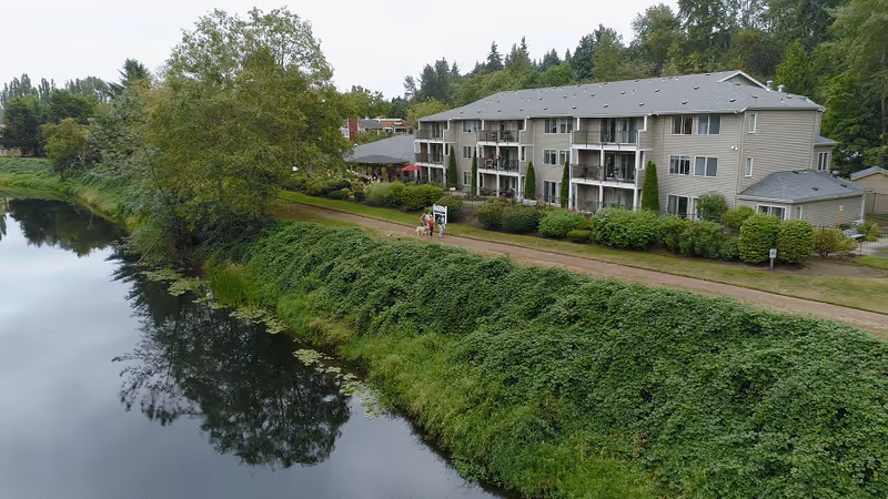 A three-story residential building with balconies overlooking a calm river, surrounded by lush greenery and trees, with a walking path along the riverbank where two people are walking.