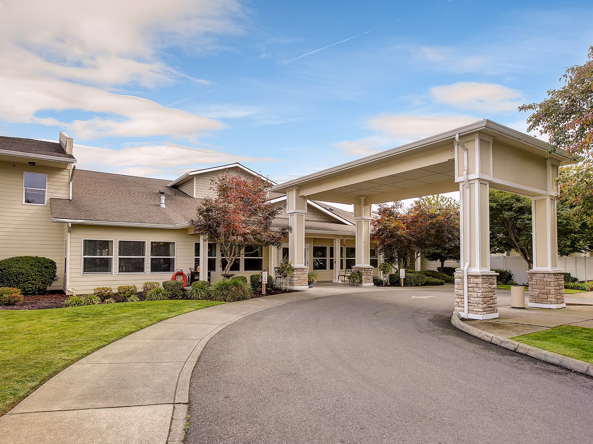 Exterior view of Prestige Senior Living Auburn Meadows showing the main entrance with a covered driveway, surrounded by well-maintained landscaping including green grass, bushes, and trees under a partly cloudy sky.