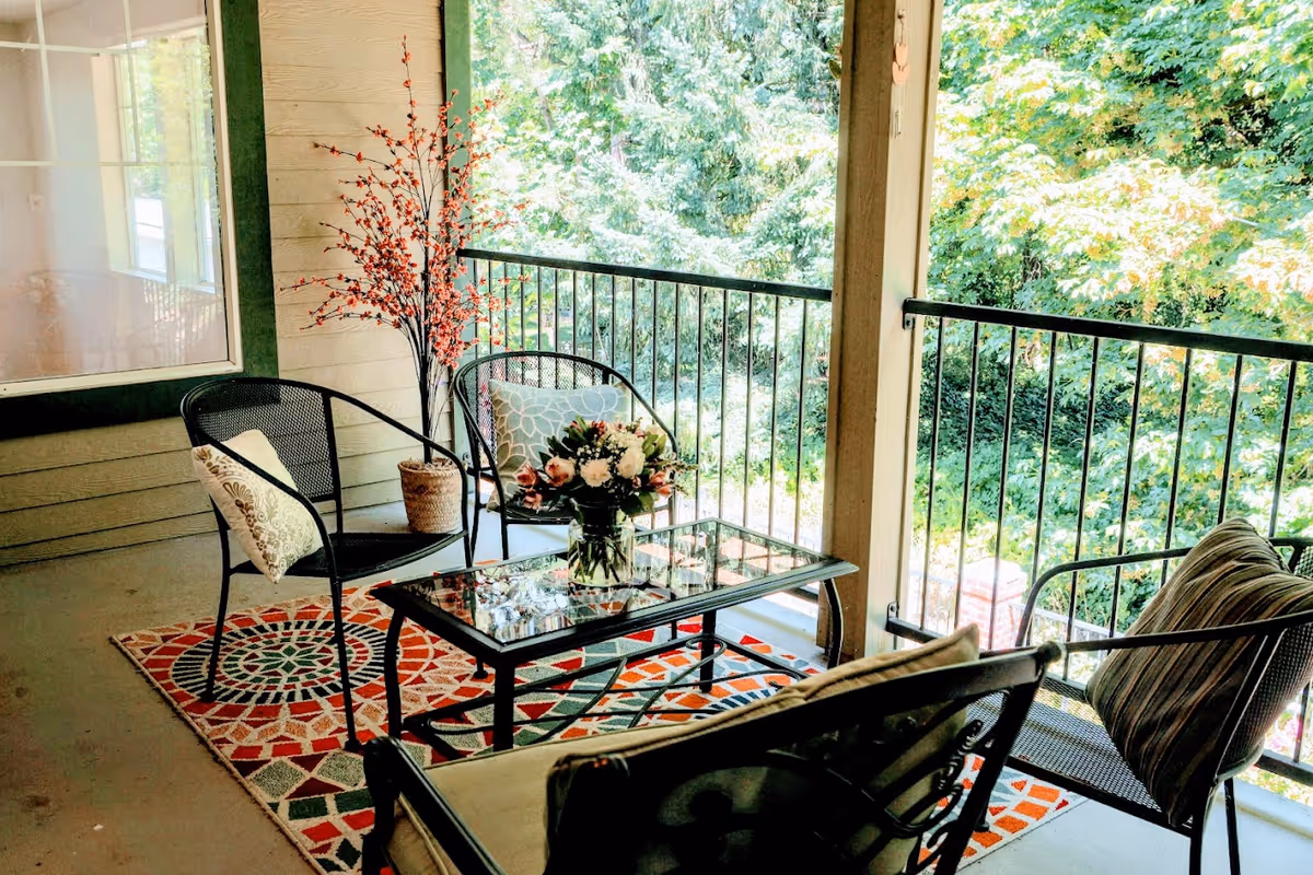 Covered balcony with metal patio chairs, a glass-top coffee table on a patterned rug, potted plants, and a leafy tree view.