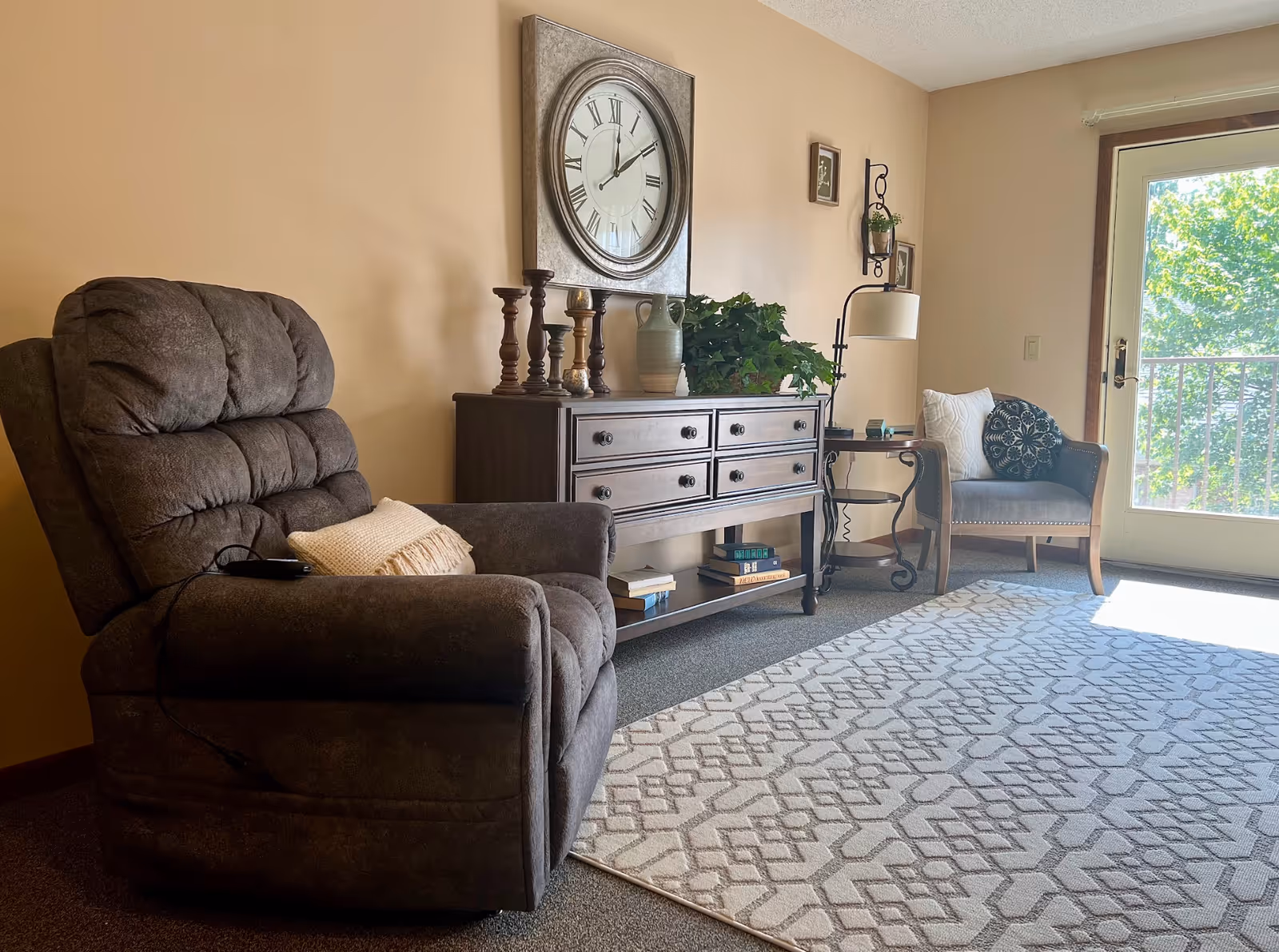 A cozy living room area with a large brown recliner chair on the left, a wooden sideboard with decorative candlesticks, a vase, and a plant in the center, and a cushioned armchair with pillows near a glass door letting in natural light. A patterned rug covers part of the carpeted floor.