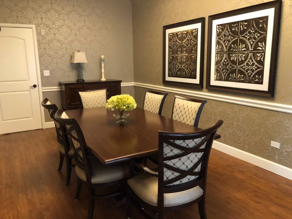 A formal dining room with a dark wooden rectangular table surrounded by eight cushioned chairs. The walls have patterned wallpaper with two framed decorative metal art pieces hanging. A sideboard with a lamp and a candle holder is against the wall, and the floor is wooden.