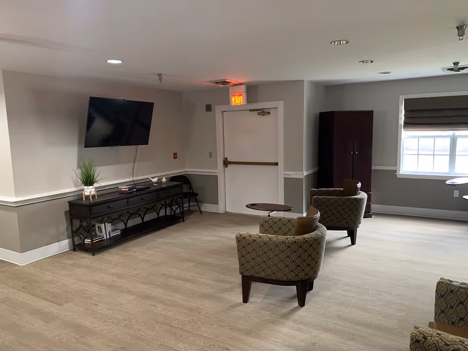 A common area with light wood flooring, two patterned armchairs facing a small round table, a dark wood cabinet, a wall-mounted flat screen TV above a console table with books and decorative items, and a window with a brown Roman shade.