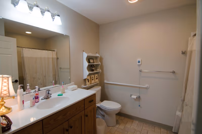 A bathroom with a wooden vanity topped with a white sink and various toiletries, a large mirror above the sink, a toilet with a white shelf above it holding decorative items, a grab bar on the wall next to the toilet, and a shower with a curtain on the right side.