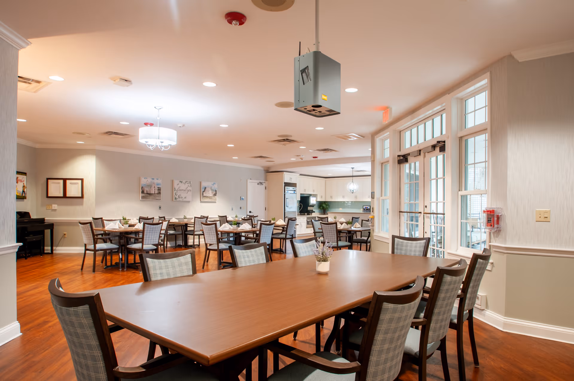 A spacious dining room in a senior living facility with multiple wooden tables and cushioned chairs arranged neatly. The room has warm wooden flooring, soft lighting from ceiling fixtures, and large windows letting in natural light. The background shows a kitchen area with white cabinets and modern appliances. Walls are decorated with framed pictures and a piano is visible on the left side.