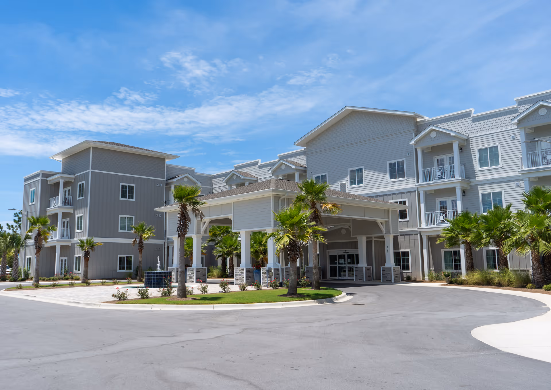 Exterior view of Seagrass Village Independent Living facility showing a large, modern three-story building with balconies, a covered entrance, palm trees, and a clear blue sky.