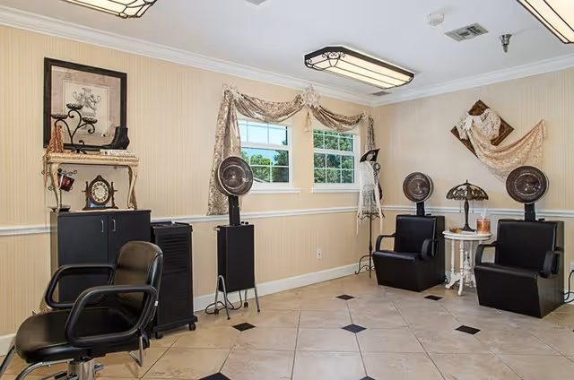 Interior of a hair salon area with three black salon chairs, two vintage hair dryers on stands, a small white table with a lamp and candle, decorative curtains on two windows, and wall decorations including a framed picture and a dress form with lace fabric.