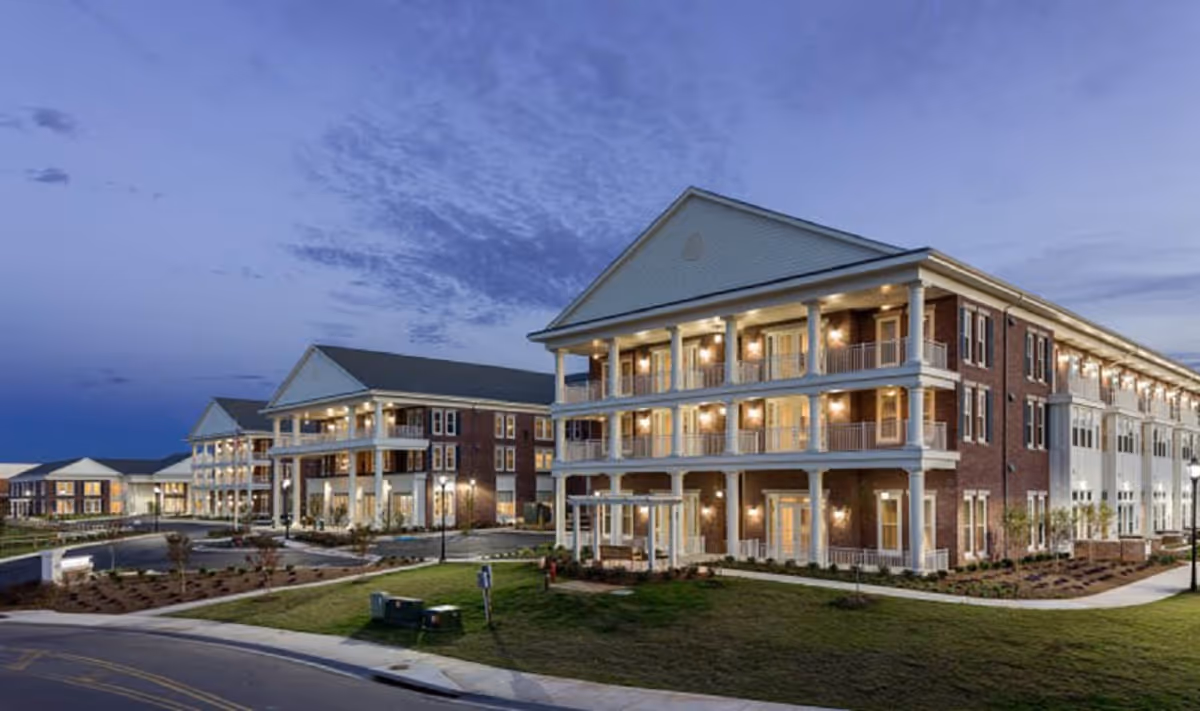 Exterior view of a large, three-story senior living facility building at dusk with multiple balconies and warm exterior lighting. The building has a brick facade with white columns and trim, surrounded by landscaped grounds and a curved driveway.