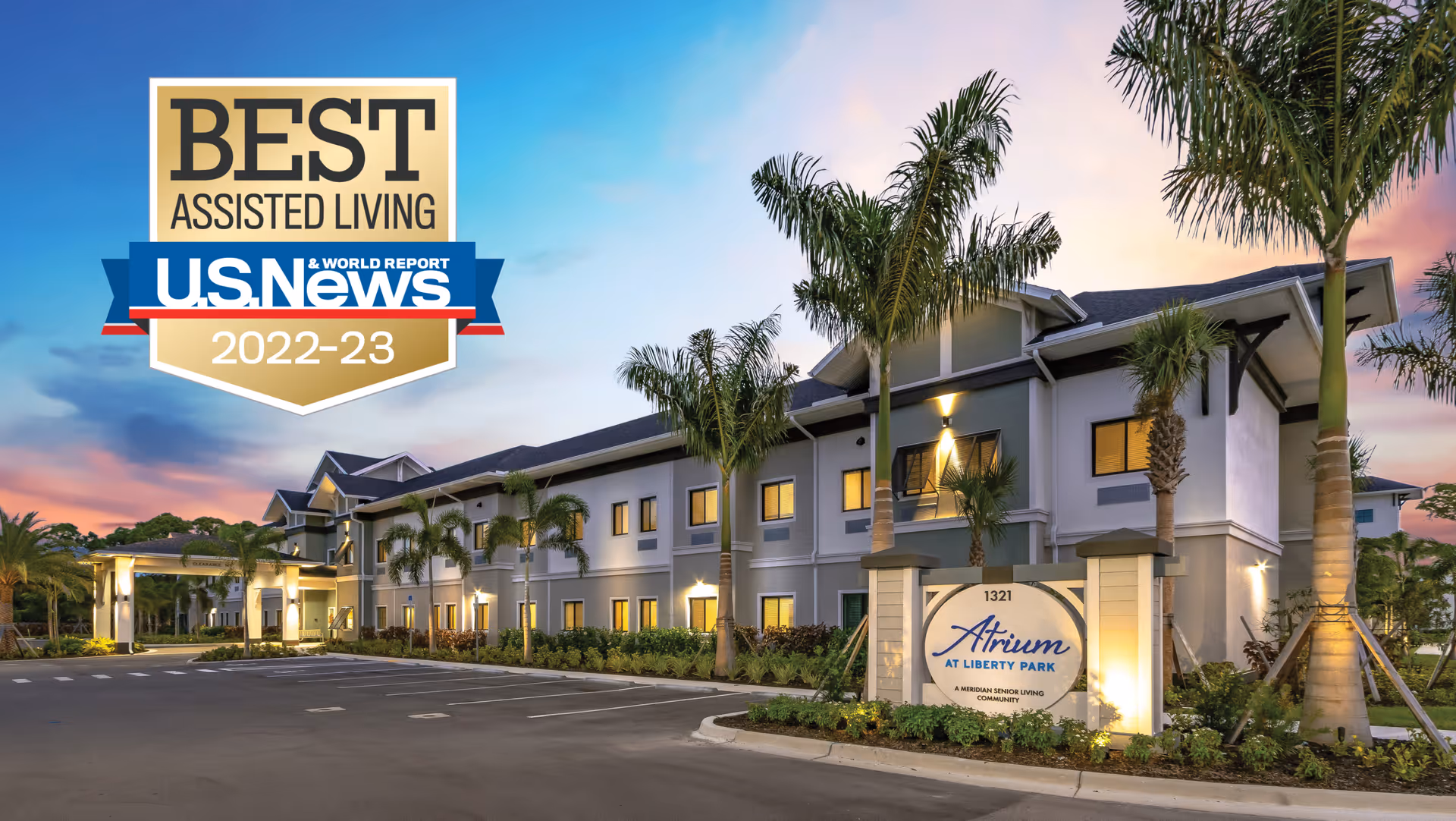 Exterior view of Atrium at Liberty Park senior living facility at sunset with palm trees and a well-lit entrance. A sign in front displays the facility name and address. A badge in the sky area indicates it was awarded Best Assisted Living by U.S. News & World Report for 2022-23.