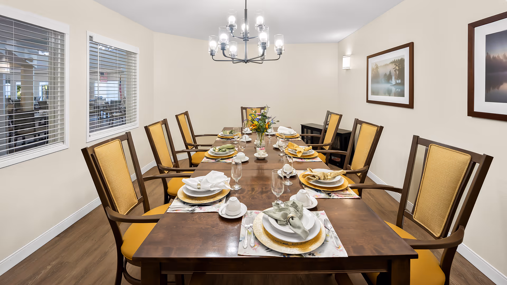 A formal dining room with a long wooden table set for eight people. Each place setting includes a plate, napkin, glass, and cup. The chairs have wooden frames with yellow cushions. The room has light-colored walls, two framed landscape pictures, and a modern chandelier hanging above the table. There are two windows with white blinds on the left wall.