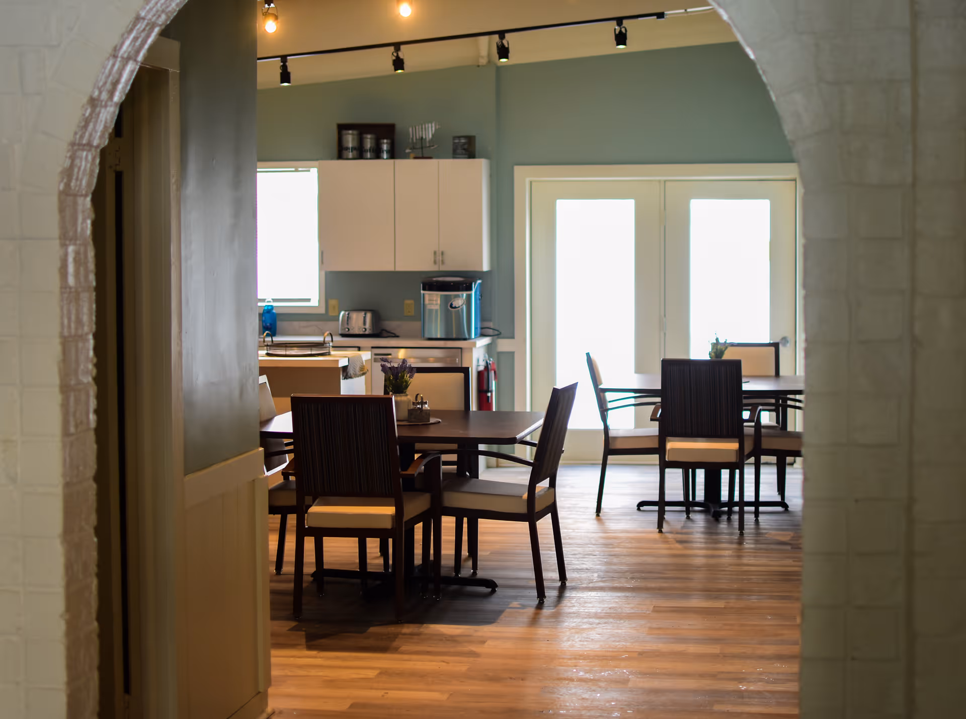View through an arched doorway into a dining area with wooden tables and chairs. The room has light blue walls, wooden flooring, and white cabinets with kitchen appliances visible in the background. Large windows and glass doors allow natural light to fill the space.