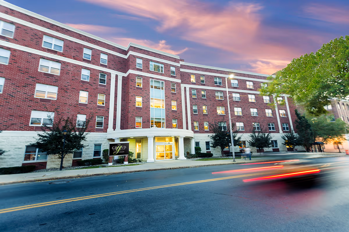Exterior view of a large brick building with multiple windows and a white stone base, identified as The Grand Rehabilitation and Nursing at River Valley, taken at dusk with a colorful sky and blurred car lights on the street in front.