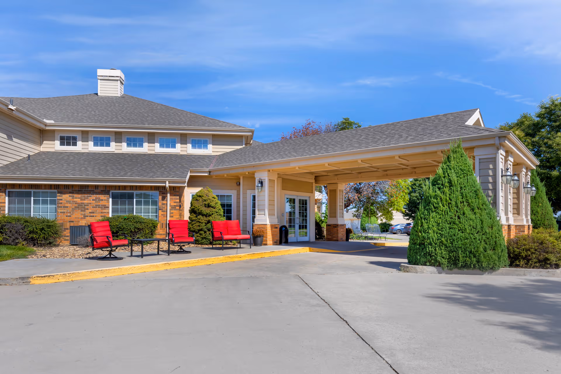 Exterior view of Brookdale Liberal Springs facility entrance with a covered drop-off area, red outdoor chairs and tables, shrubs, and a clear blue sky.