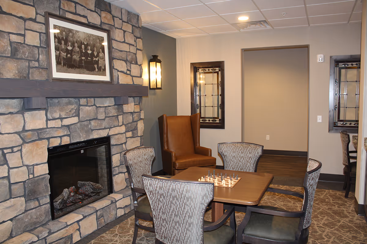 A cozy common area with a stone fireplace, a framed black and white photo above the mantel, a wooden table with a chessboard, four patterned chairs around the table, and a brown leather wingback chair against the wall.
