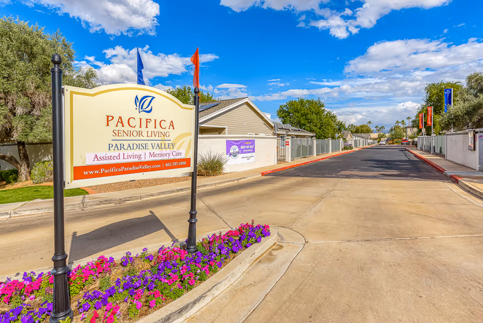 Entrance sign for Pacifica Senior Living Paradise Valley beside a driveway lined with flowers, flags, and gated buildings under a blue sky.