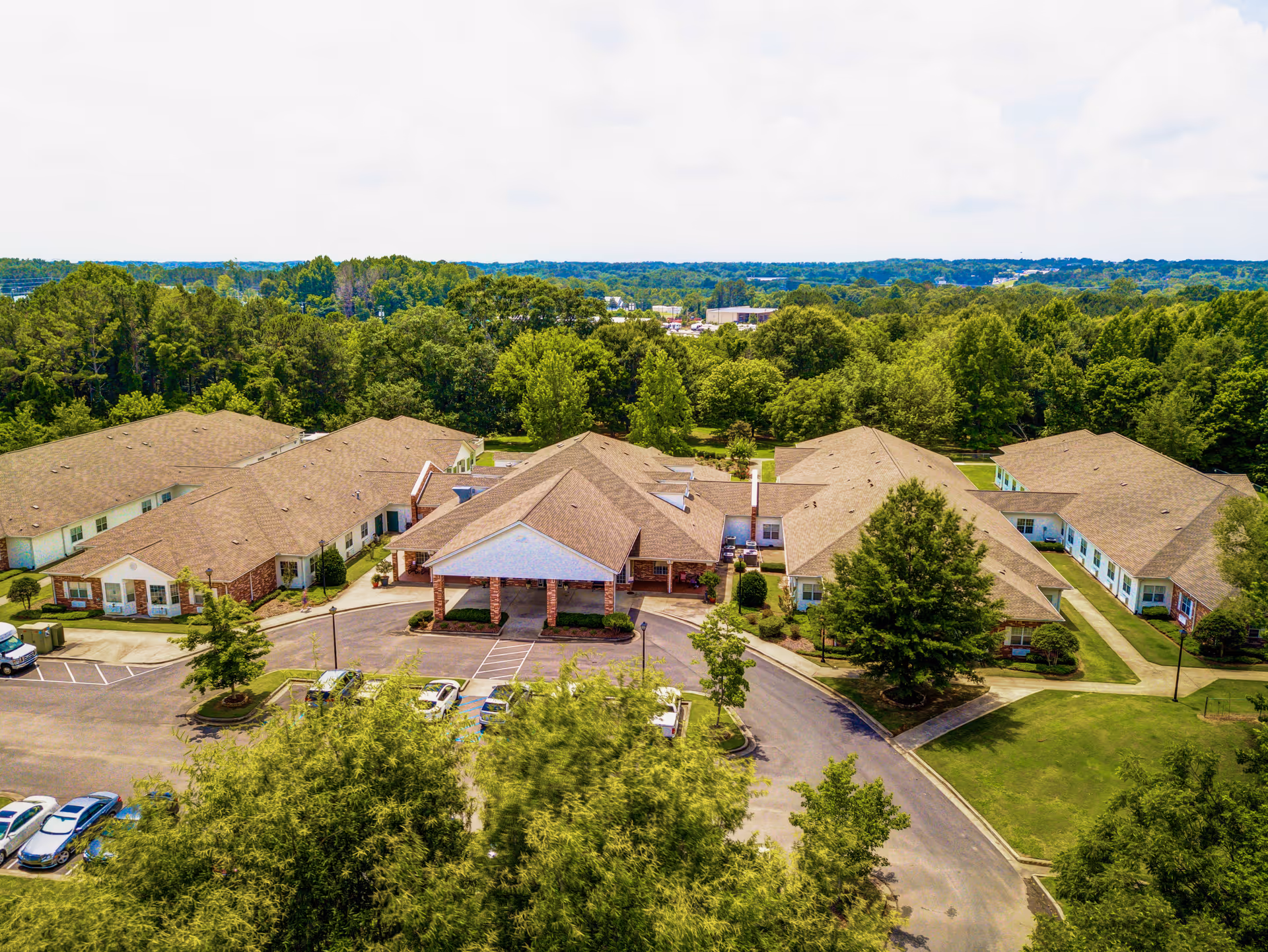 Aerial view of Bridgewood Gardens senior living facility showing multiple single-story buildings with brown roofs surrounded by green trees and a parking area with several cars.