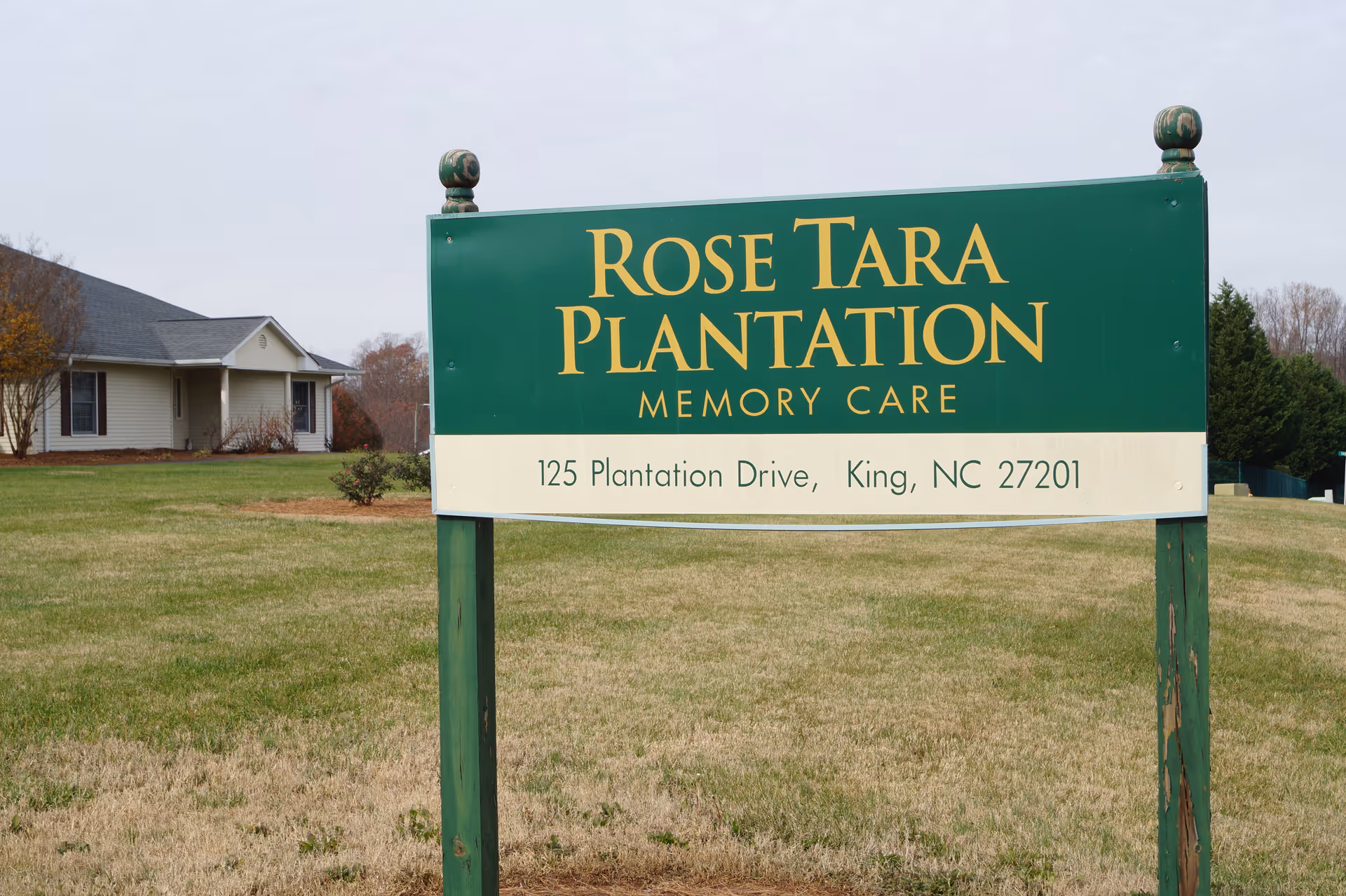 A green sign that reads "Rose Tara Plantation Memory Care" with the address, standing on a lawn in front of a single-story building.