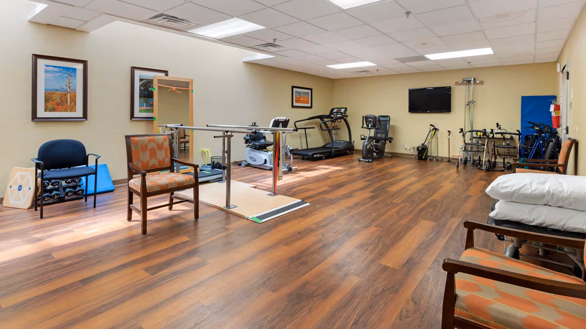 A spacious rehabilitation or exercise room with wooden flooring, featuring parallel bars for walking practice, exercise equipment including a treadmill and stationary bike, chairs, walkers, and a wall-mounted TV. The room has beige walls with framed pictures and a ceiling with fluorescent lighting.