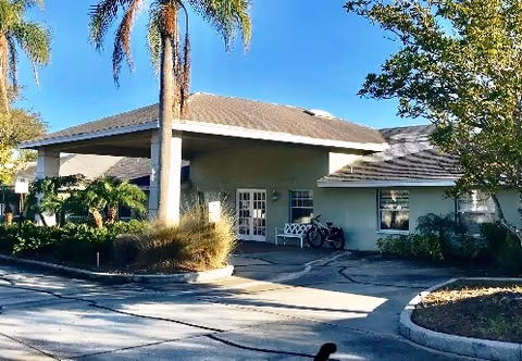 Exterior view of a single-story building with a covered entrance, surrounded by palm trees and other greenery under a clear blue sky.